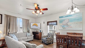 Ein Wohnzimmer mit Sofa, Sessel, Holzesstisch und Stühlen. Es gibt einen Deckenventilator, eine Lampe und ein Gemälde mit Strandmotiv.