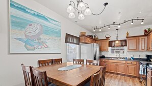 Ein Essbereich mit Holztisch und -stühlen, eine Küche mit Holzschränken und einem Kühlschrank sowie ein Meeresraubild an der Wand.