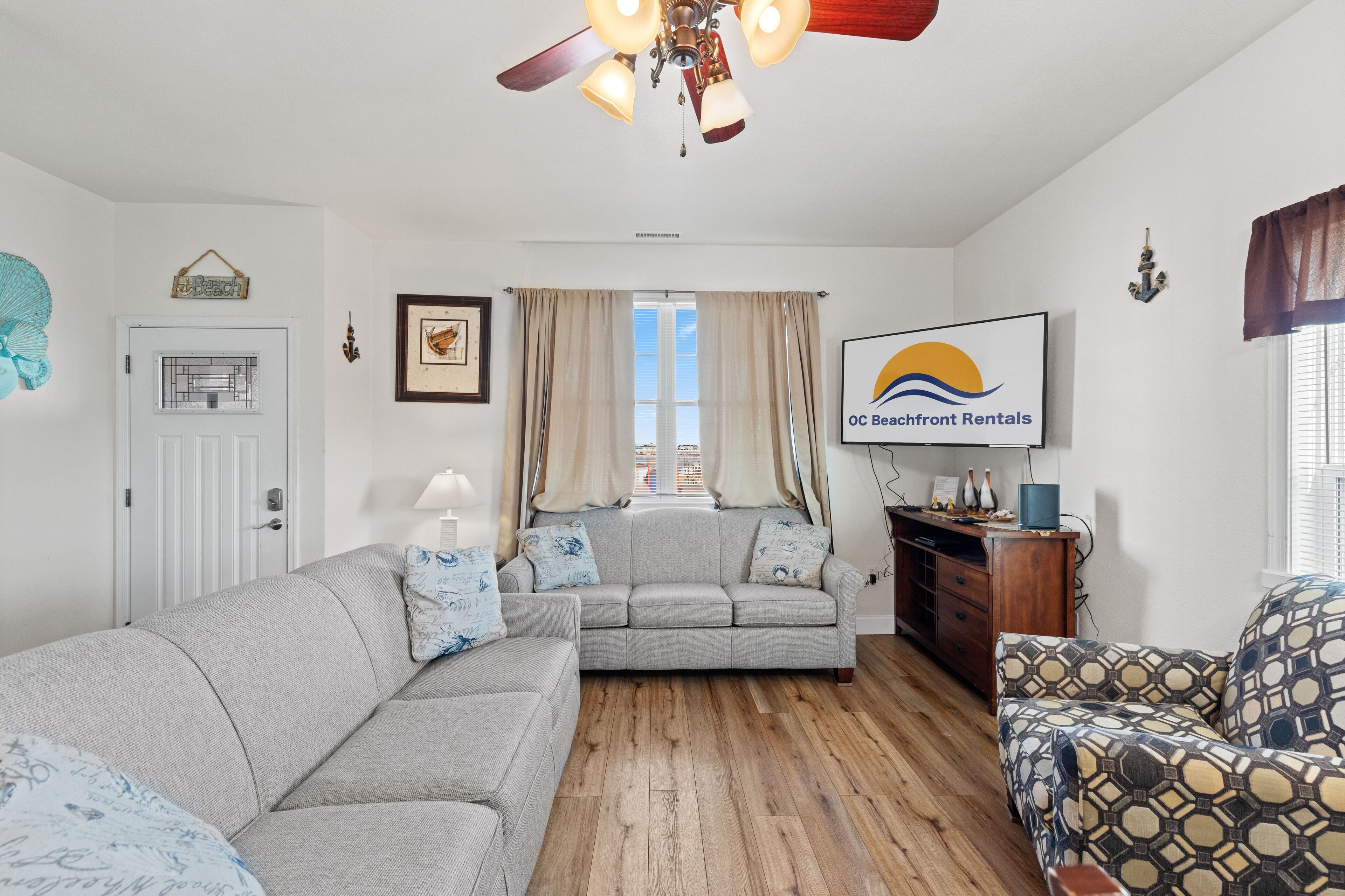 Un salon avec un canapé gris, un fauteuil à motifs, un plancher en bois et un ventilateur de plafond.