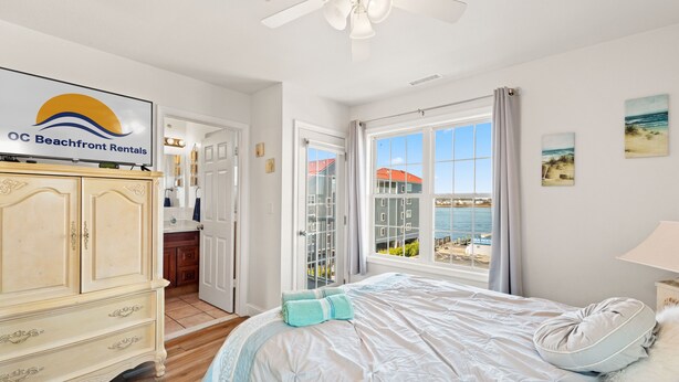 Ein Schlafzimmer mit Bett, Deckenventilator, Fernseher, der 'OC Beachfront Rentals' anzeigt, Fenster mit Meerblick und einer Tür zu einem Badezimmer.