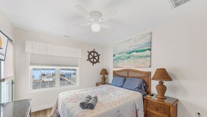 A bedroom with a bed, a window with a view of a marina, a ceiling fan, a ship's wheel decoration, and a painting of the ocean.