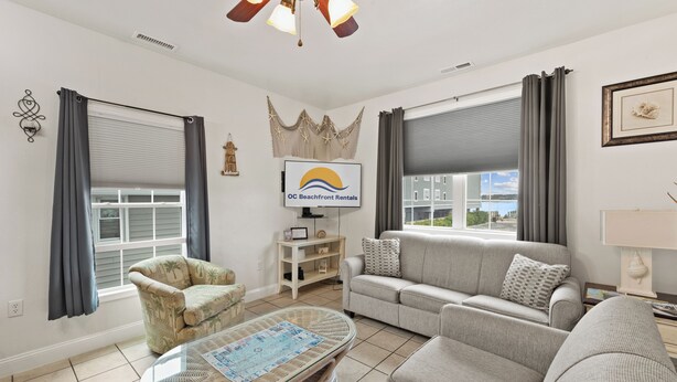 Una sala de estar con un sofá, un sillón y una mesa de centro. Hay un televisor en la pared y una ventana con cortinas.