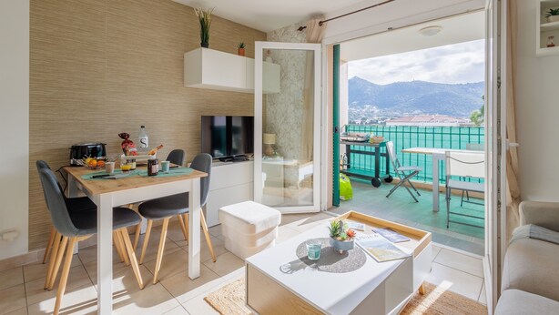 Un salon moderne comprenant un coin repas, un canapé, une table basse et un balcon offrant une vue sur la mer et les montagnes.