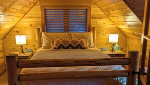 A cozy attic bedroom with a wooden bed frame, two bedside lamps, and a window with blinds. -  Cozy, charming two bedroom McKenzie Bridge River front cabin for 2 Guests, (McKenzie Bridge)