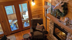 A cozy cabin interior with a fireplace, a comfortable seating area, and a view through the glass doors to the outside. -  Cozy, charming two bedroom McKenzie Bridge River front cabin for 2 Guests, (McKenzie Bridge)