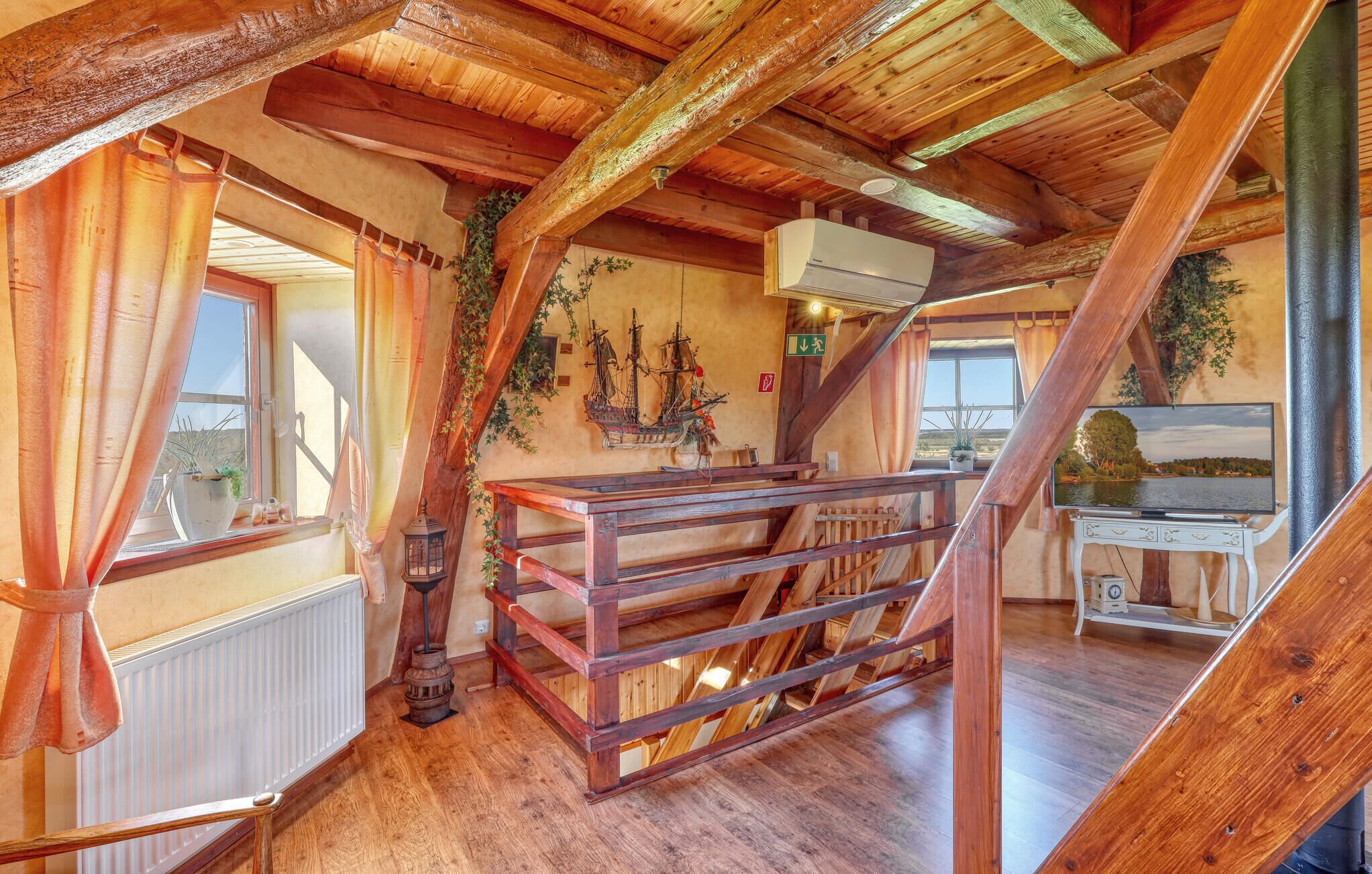Ein rustikaler Holzinnenraum mit schrägen Decken, einer Holztreppe, einem Heizkörper, einem kleinen Tisch mit einem Fernseher und einem Schiffsmodell an der Wand.