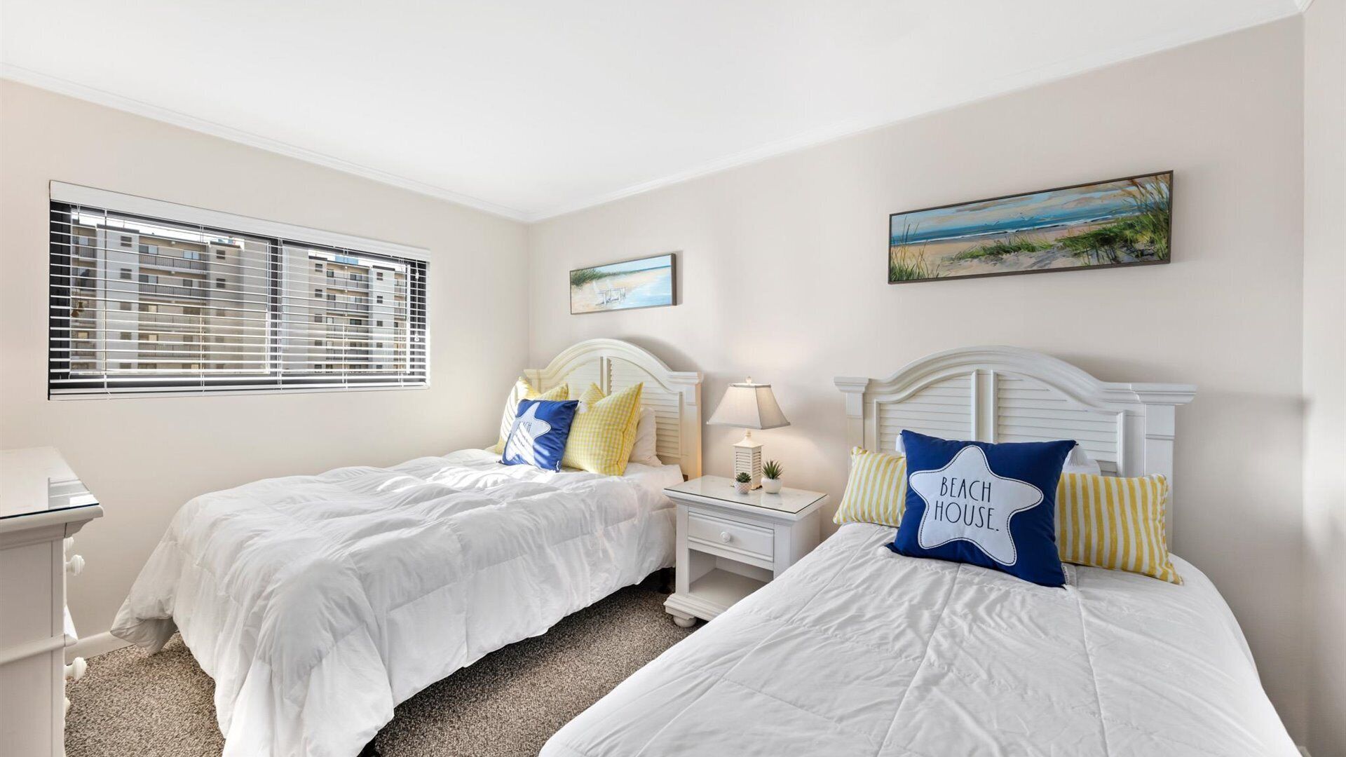 A bedroom with two beds, a nightstand with a lamp, a window with blinds, and a painting on the wall.