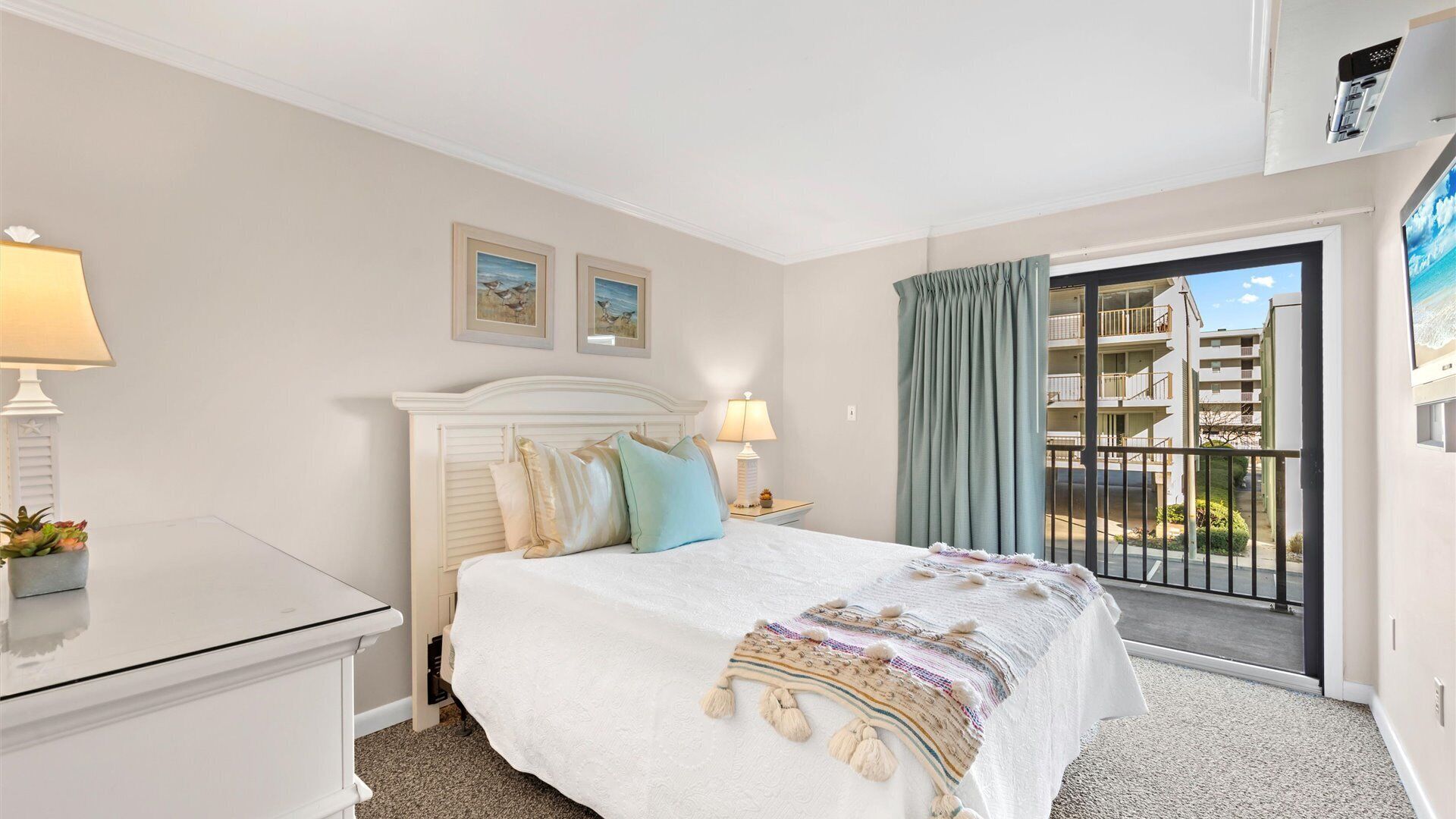 A hotel room with a bed, bedside table, lamp, and a view of a balcony with buildings.