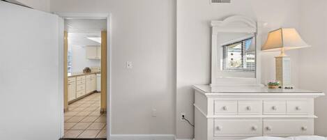 A bedroom with a white dresser, a mirror, and a lamp. There is a door leading to a bathroom and a kitchen.