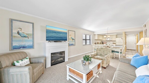 Cozy living room with fireplace, comfortable seating, and artwork on the walls, featuring a kitchen area and dining space.