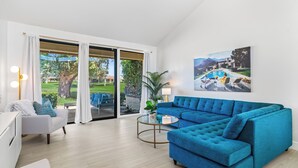 A modern living room with a blue sofa, a white armchair, and a glass coffee table. There is a large window with curtains, a painting on the wall, and a view of a pool and greenery outside.
