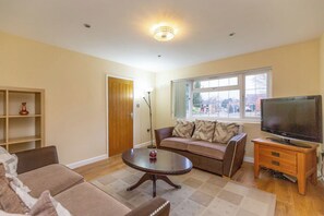 Una sala de estar con un sofá, una mesa de centro, un televisor sobre un mueble de madera, una ventana con vistas al exterior y una puerta.