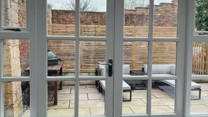 Vista a través de un ventanal hacia un patio con pared de ladrillo, valla de madera y mobiliario exterior.