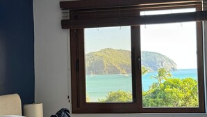 A bedroom with a large window overlooking a coastal landscape, a bed with multiple pillows, and a ceiling fan. - San Juan del Sur most exclusive condo! (San Juan del Sur)