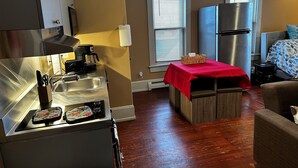 A compact kitchen with a stove, sink, and refrigerator. There is a small dining table with a red tablecloth and a basket. The room has wooden flooring and a window with blinds. - Apt 2B Lake Bomoseen Inn “The Dog” with boat slip (Castleton)