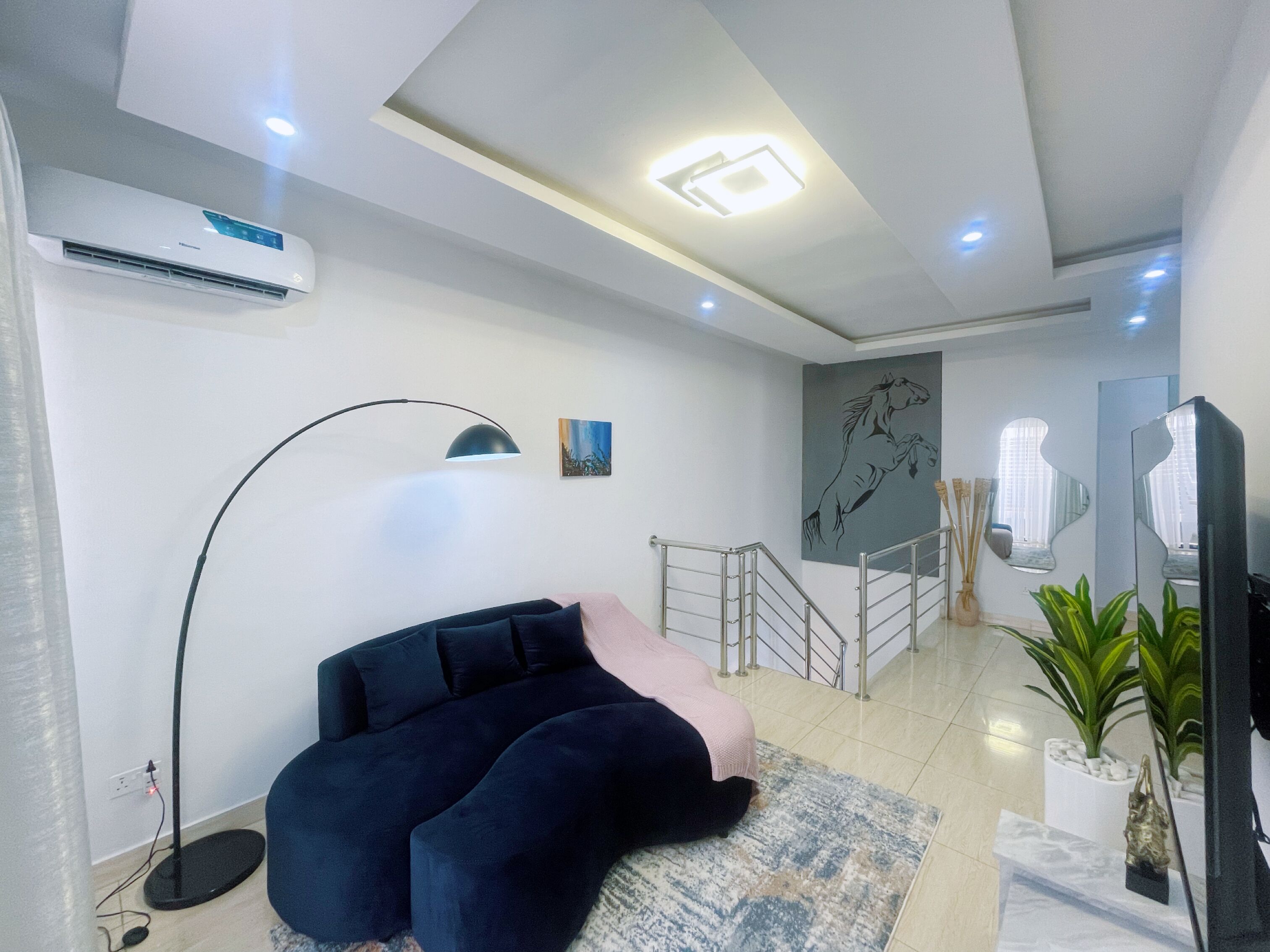 A modern living room with a dark blue sofa, a floor lamp, a painting, and a staircase leading to another level.