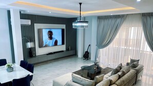 A modern living room with a large flat-screen TV, a sofa, a dining table with chairs, and a guitar.