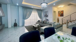 A modern living room with a sofa, coffee table, and dining area. There is a staircase and a chandelier.