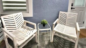 Una habitación con dos sillas blancas, una mesita pequeña, una planta en maceta y una ventana con persianas.