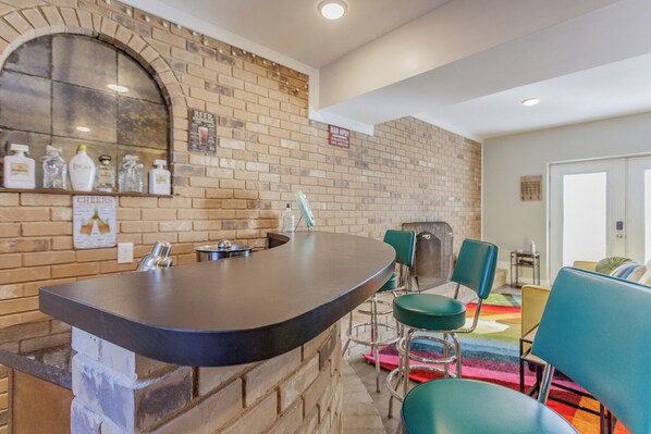 A bar area with a brick wall, bar stools, and a counter. There are bottles and glasses on the wall, and a sign that says "Cheers".