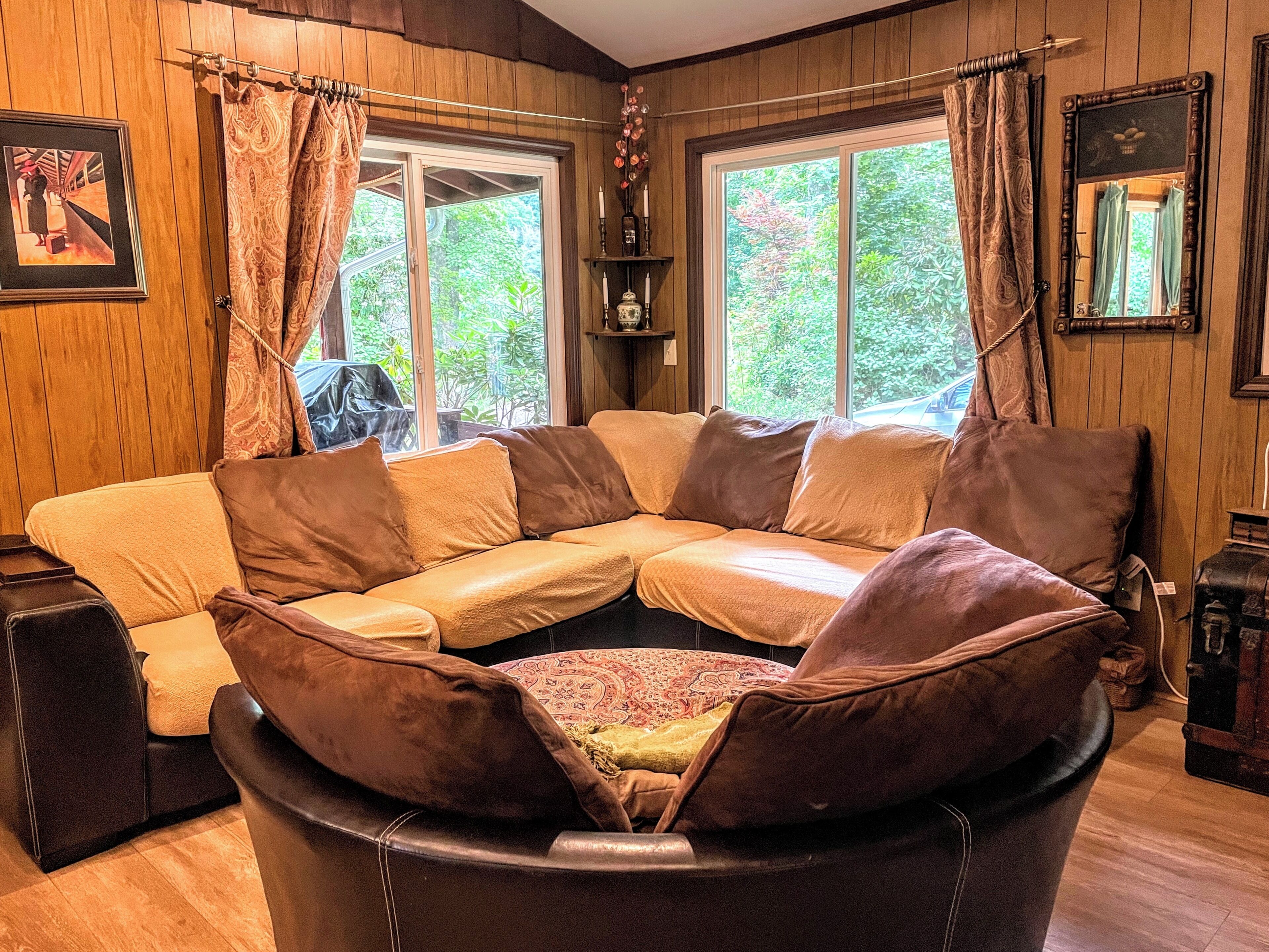 Un salón con esquina, con un sofá de cuero marrón, piso de madera y una ventana con cortinas.