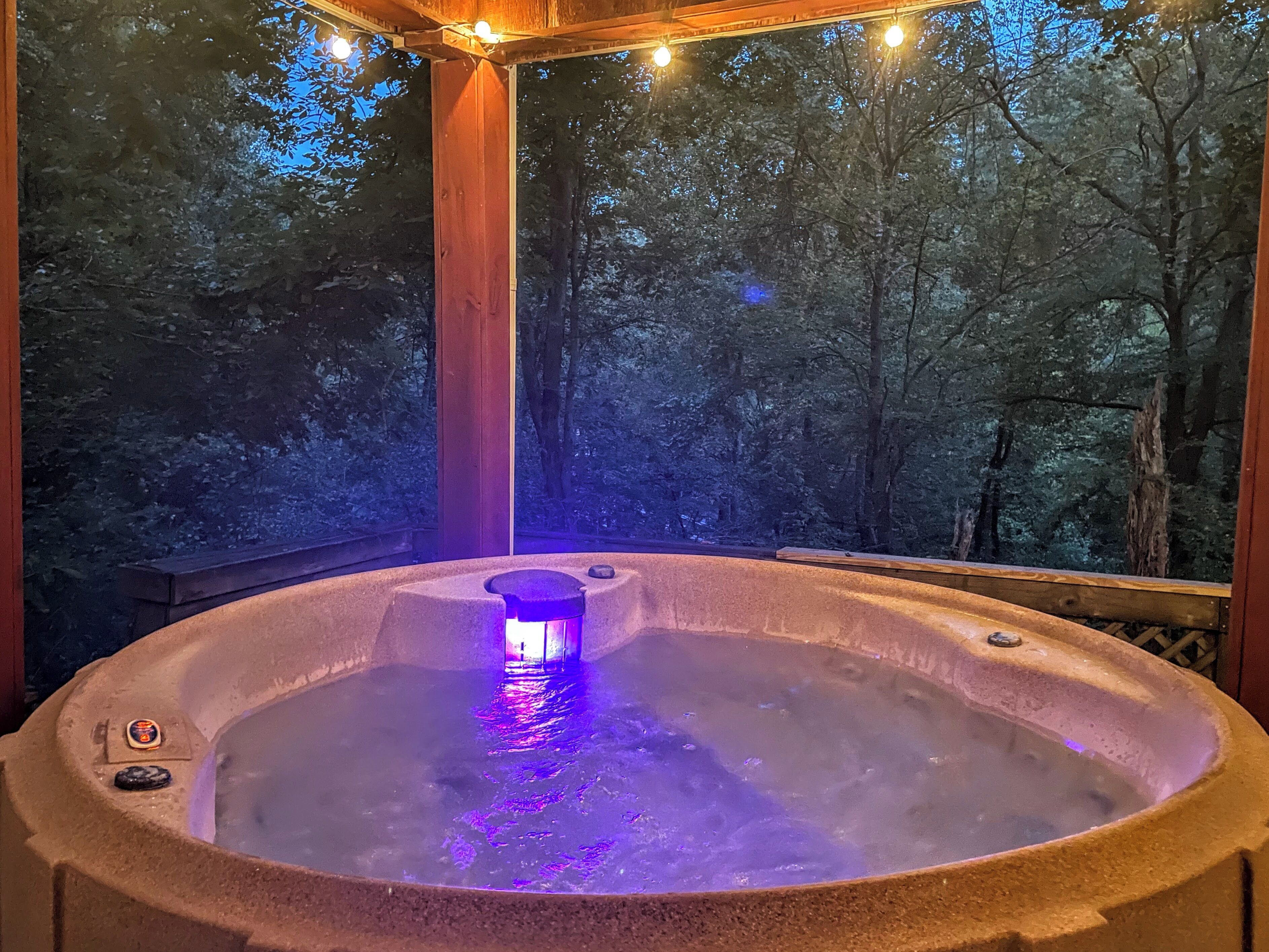A hot tub with a built-in light, surrounded by a wooden deck and a forested backdrop.