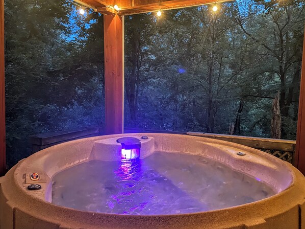 A hot tub with a built-in light, surrounded by a wooden deck and a forested backdrop.