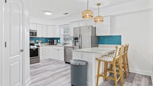 Una cocina moderna con armarios blancos, un refrigerador de acero inoxidable, un microondas y un lavamanos. Hay tres lámparas colgantes y un bote de basura.