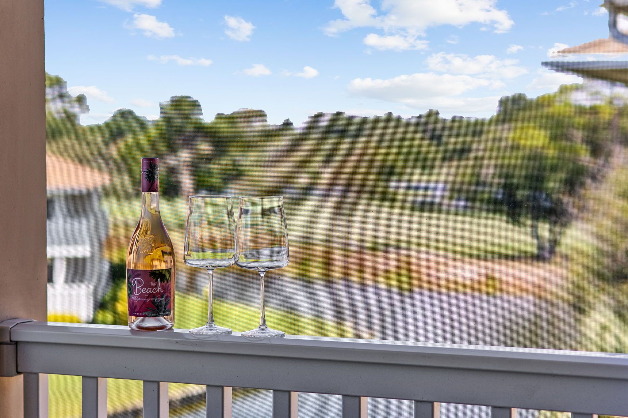 A bottle of wine and two glasses on a balcony railing with a view of a golf course.