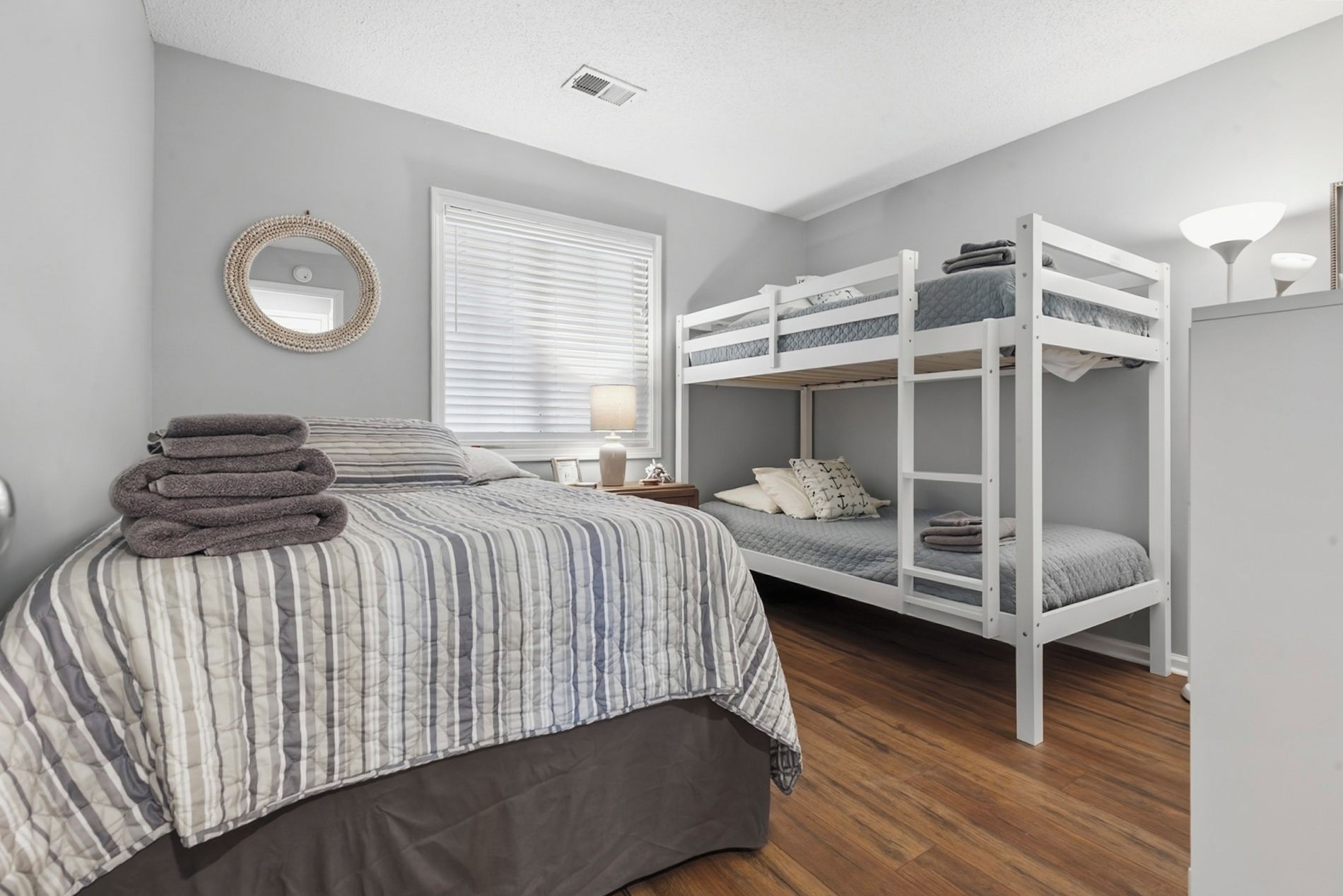 A bedroom with a bunk bed, a single bed, a bedside table, a lamp, a mirror, and a window with blinds.