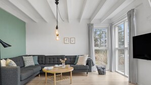 Een moderne woonkamer met een grijze modulaire bank, een ronde houten salontafel en een zwarte vloerlamp. Er is een groot raam met gordijnen, een flatscreen televisie aan de muur bevestigd en ingelijste foto’s.