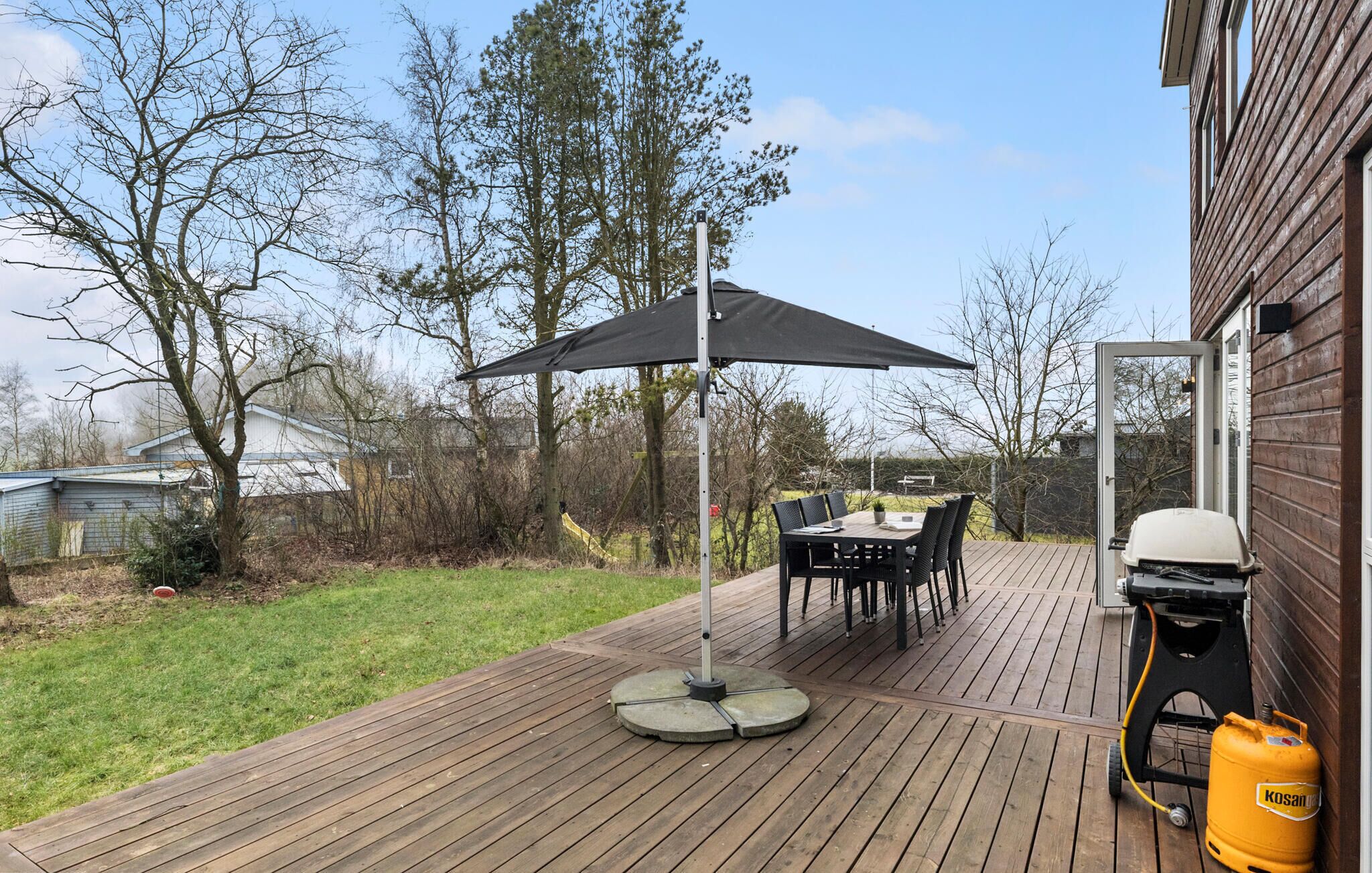 Eine Holzterrasse mit Tisch und Stühlen unter einem Sonnensegel, ein Grill und ein Gasbehälter.