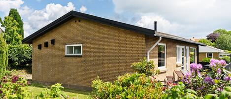 Et etplans murhus med et skråt tag, en lille veranda med en stol, og en have med forskellige planter og blomster.