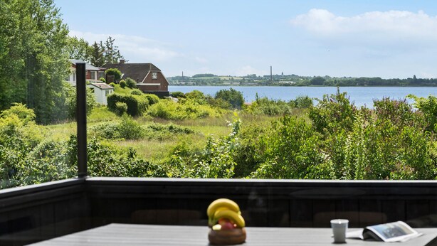 A deck with a wooden table, a banana, and a cup. In the background, there is a house by a lake with a clear sky. - 4 bedroom gorgeous home in Gråsten (Gråsten)