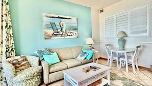 Een woonkamer met een blauwgroene muur, een beige bank, een fauteuil met een patroon, een salontafel, een eettafel met stoelen en een schilderij van een strandlandschap.