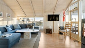 A modern living room with a blue sofa, a round coffee table, and a wooden chair. A flat-screen TV is mounted on the wall, and a flag hangs beside it. - Gorgeous home in Juelsminde with sauna (Juelsminde)