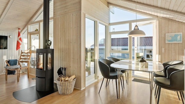 A modern interior with a wood-burning stove, a dining area with chairs, and large windows offering a view of the outside. - Gorgeous home in Juelsminde with sauna (Juelsminde)