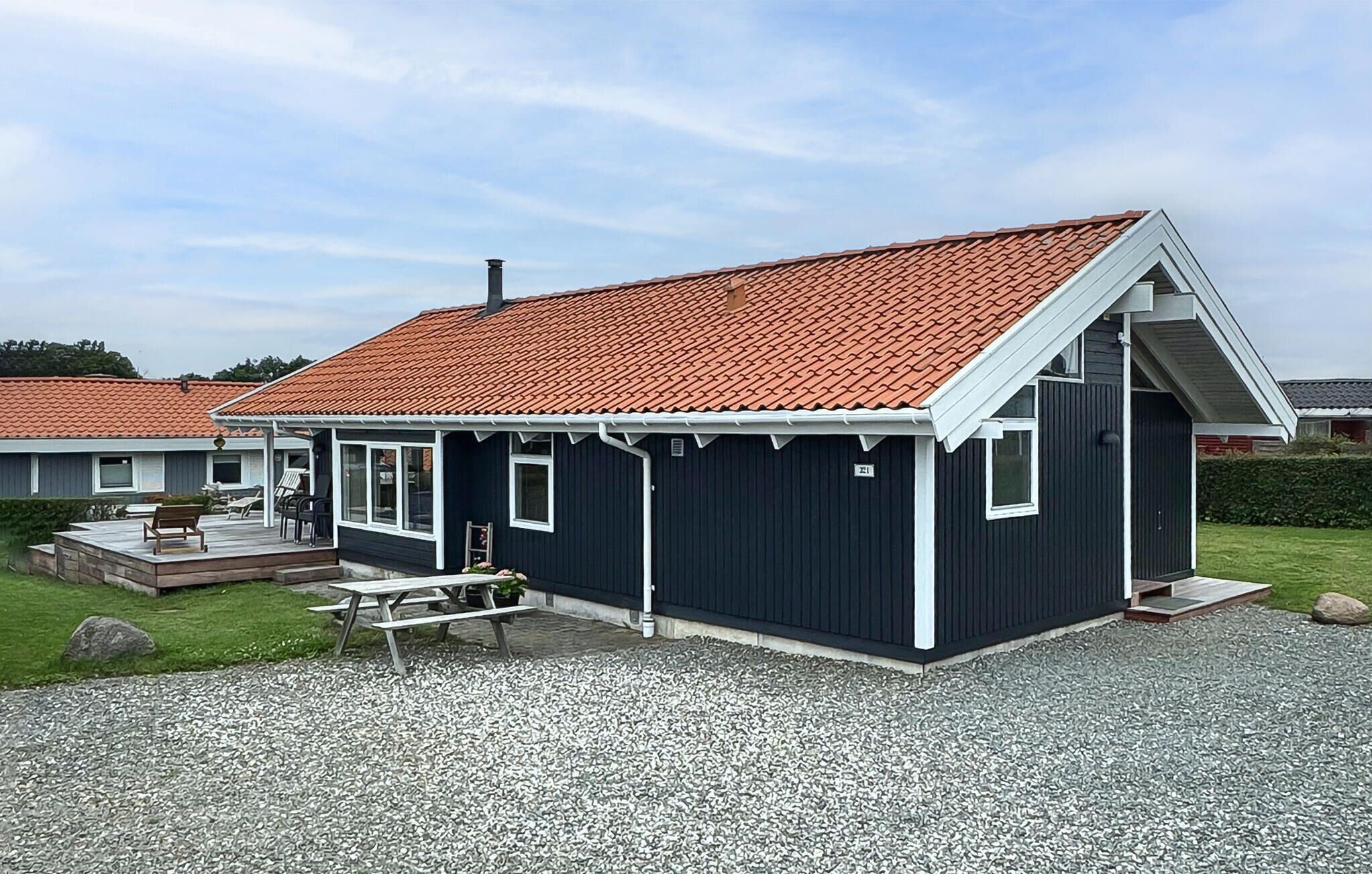 Et lille, mørkt hus med røde tegltage, en veranda med siddepladser og en grusbelagt indkørsel.