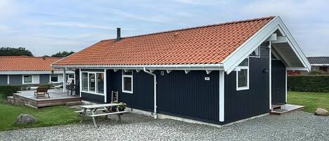Et lille, mørkt hus med røde tegltage, en veranda med siddepladser og en grusbelagt indkørsel.