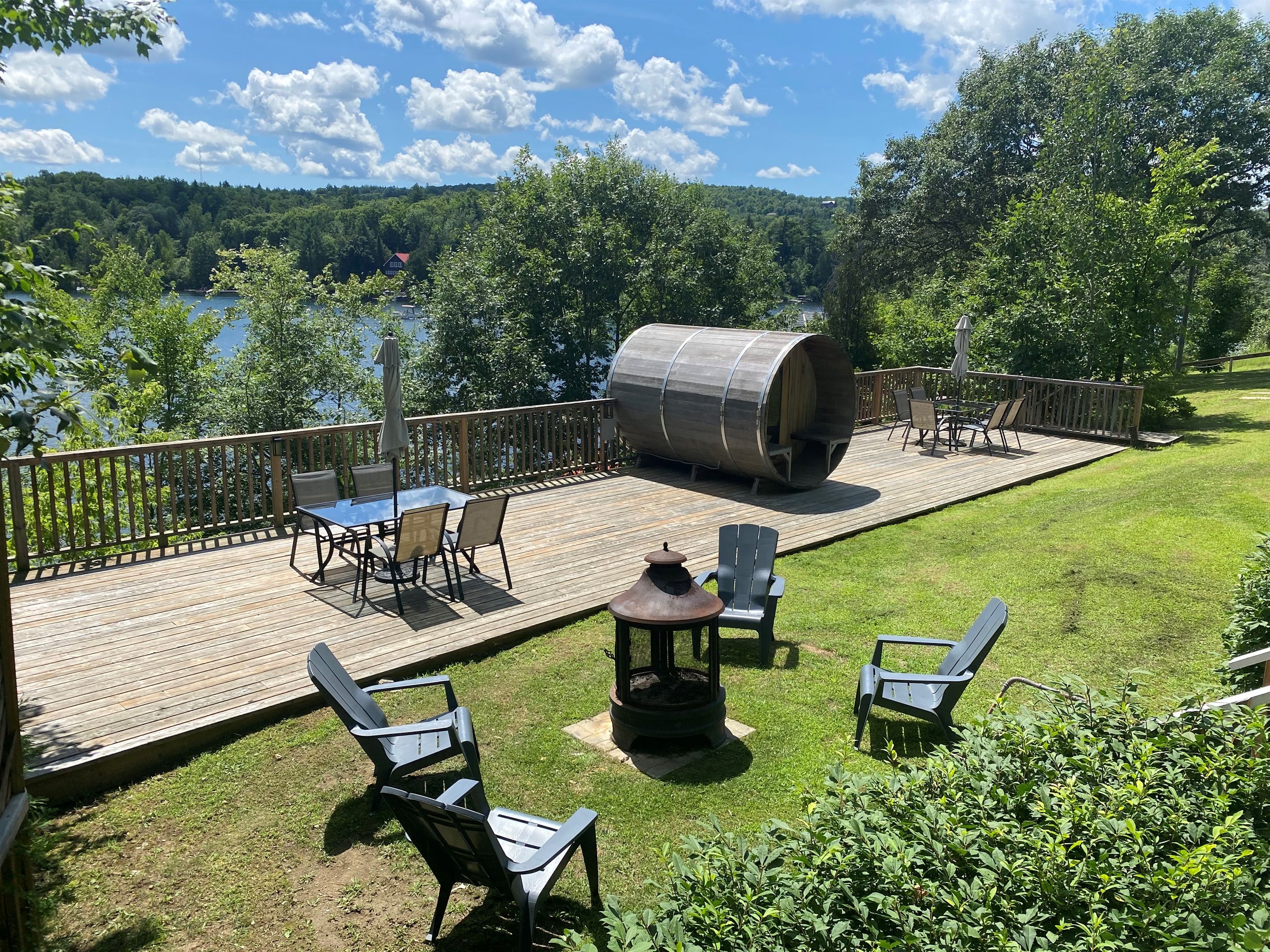 A deck with outdoor furniture, a metal barrel, and a view of a lake and forest.