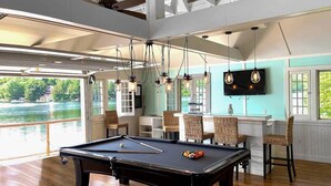 Une table de billard dans une pièce au plancher en bois, avec des chaises en osier et une vue sur un lac.