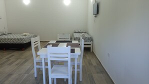 Una habitación pequeña y bien iluminada con dos camas, una mesa de comedor y sillas. Cuenta con un ventilador de techo y un televisor montado en la pared.