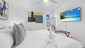 Habitación de hotel con una cama grande, ventilador de techo, televisor mostrando una escena de playa y vistas a la playa por la ventana.