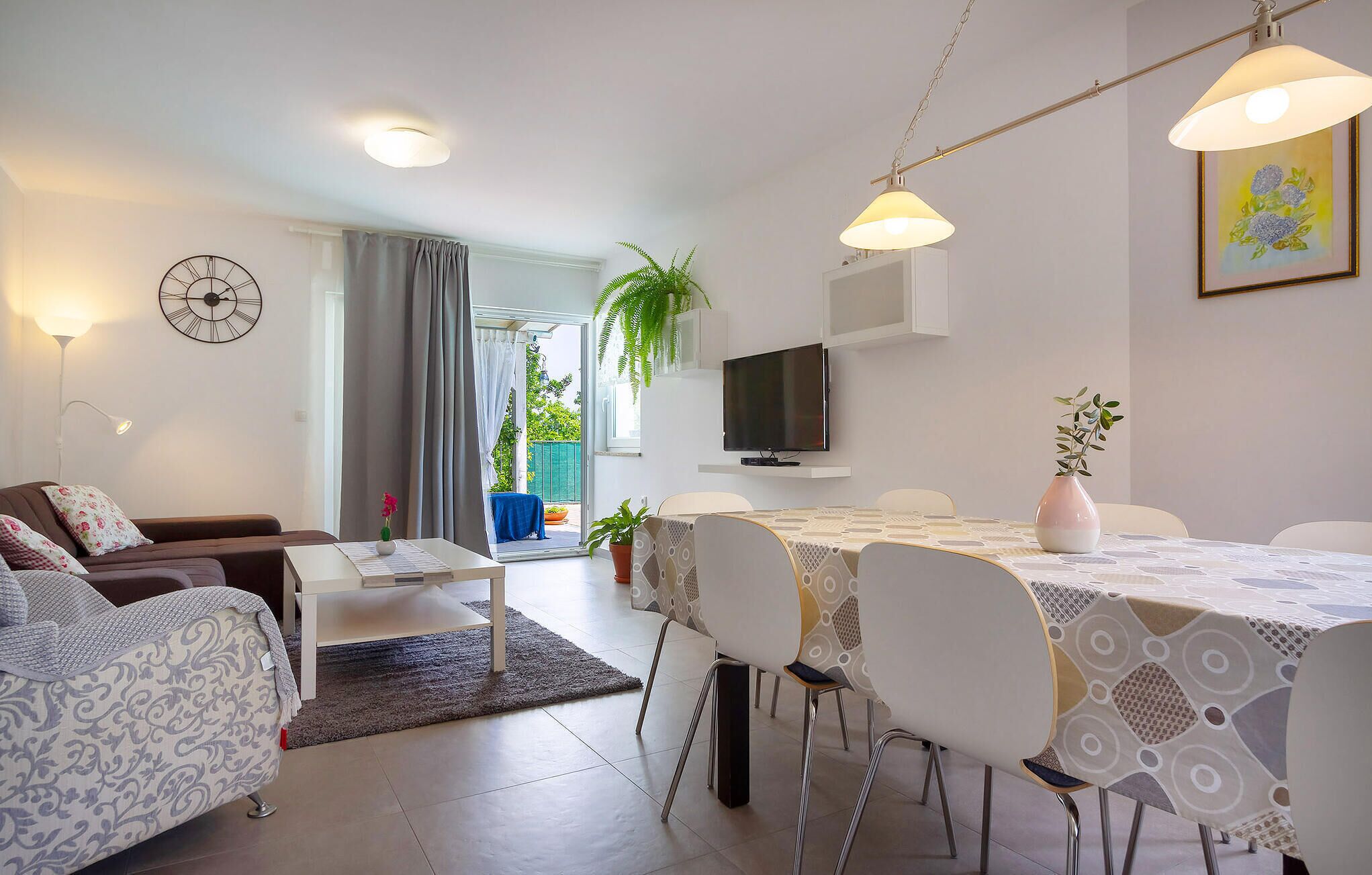 Ein modernes Wohnzimmer mit einer Couch, einem Esstisch mit Stühlen, einem Fernseher und einem großen Fenster mit Vorhängen.