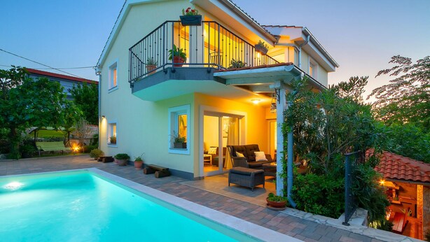 Una casa de dos pisos con piscina, mobiliario exterior y un balcón con plantas en macetas.