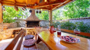 Un área de comedor al aire libre con madera, una chimenea de piedra, bancos y una mesa dispuesta con comida y copas de vino.