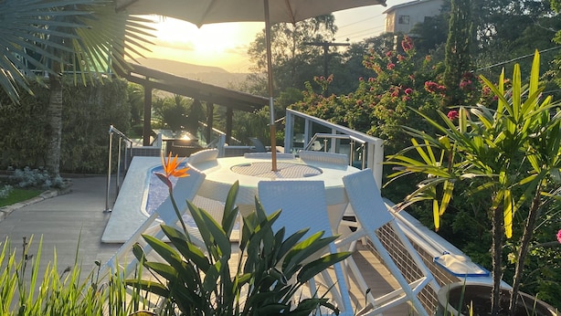 Een zonnig terras met een grote parasol, omringd door weelderige begroeiing en bloeiende planten.