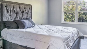 Ein Schlafzimmer mit einer gesteppten Bettdecke, einem großen Bett mit einem rautenförmigen Kopfstück und einem Fenster mit Blick auf Bäume.