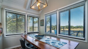 A dining area with a wooden table, chairs, and a bowl on a placemat. There are large windows with blinds, and a ceiling light fixture is visible. - 2BR/2BA + Loft Cottage! Close to Beach! Golf Course Views! Amenity access Available! (Seabrook Island)