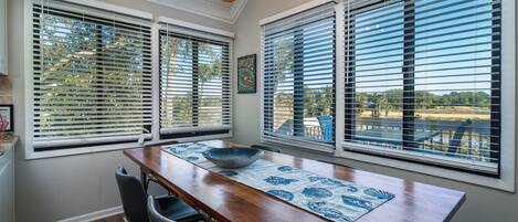 A dining area with a wooden table, chairs, and a bowl on a placemat. There are large windows with blinds, and a ceiling light fixture is visible.